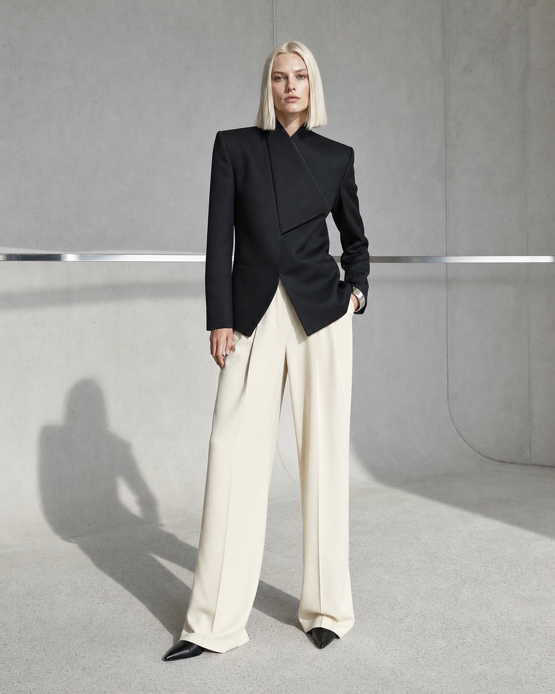 Fictional blonde fashion model in sculptural tailoring inside a concrete studio